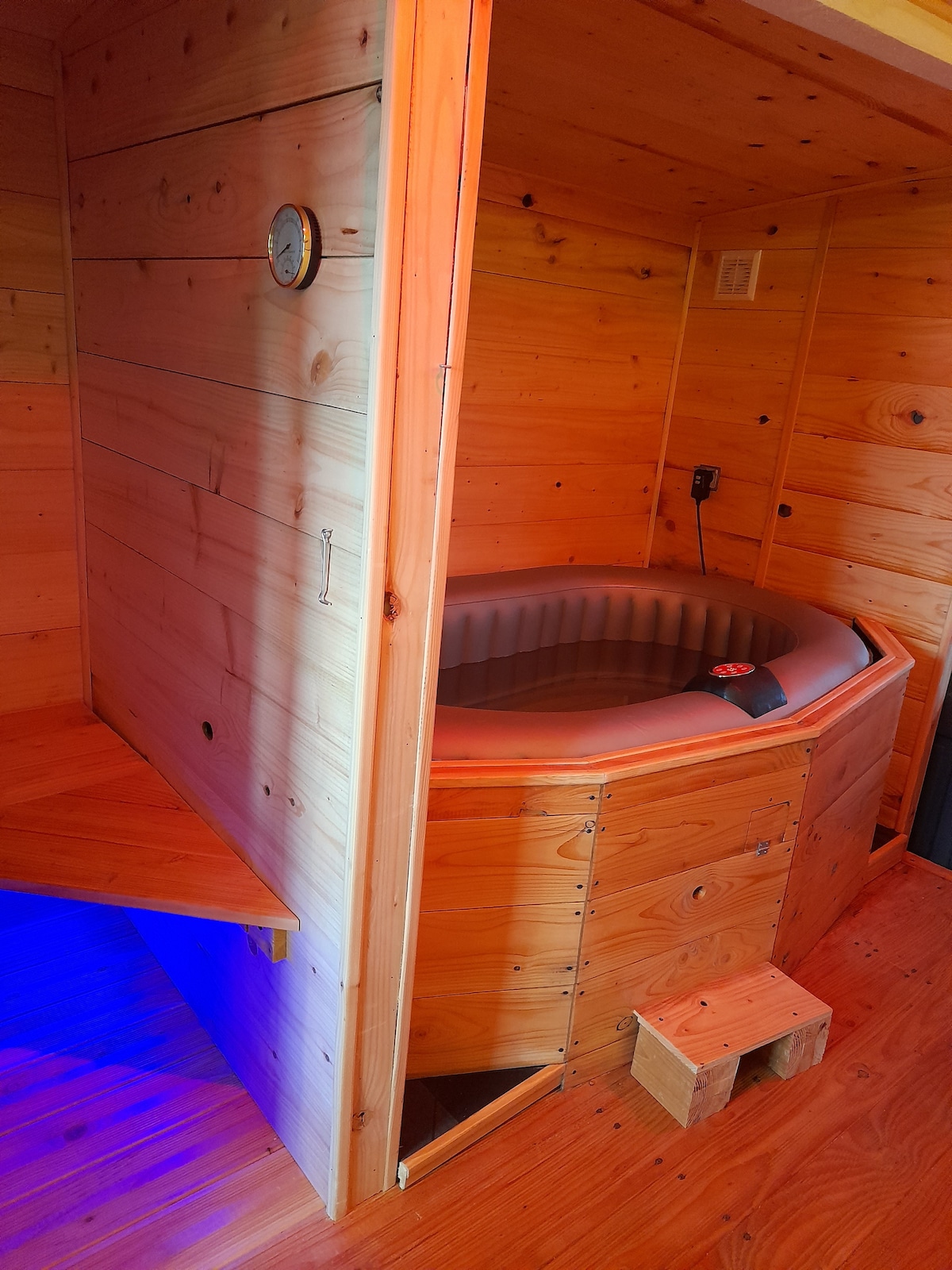 A wooden-accented space showcases a hot tub set into the corner, surrounded by warm-toned walls and flooring. A small shelf to the side offers a resting area, while a wall clock provides an element of timekeeping. Soft lighting adds to the relaxed atmosphere.