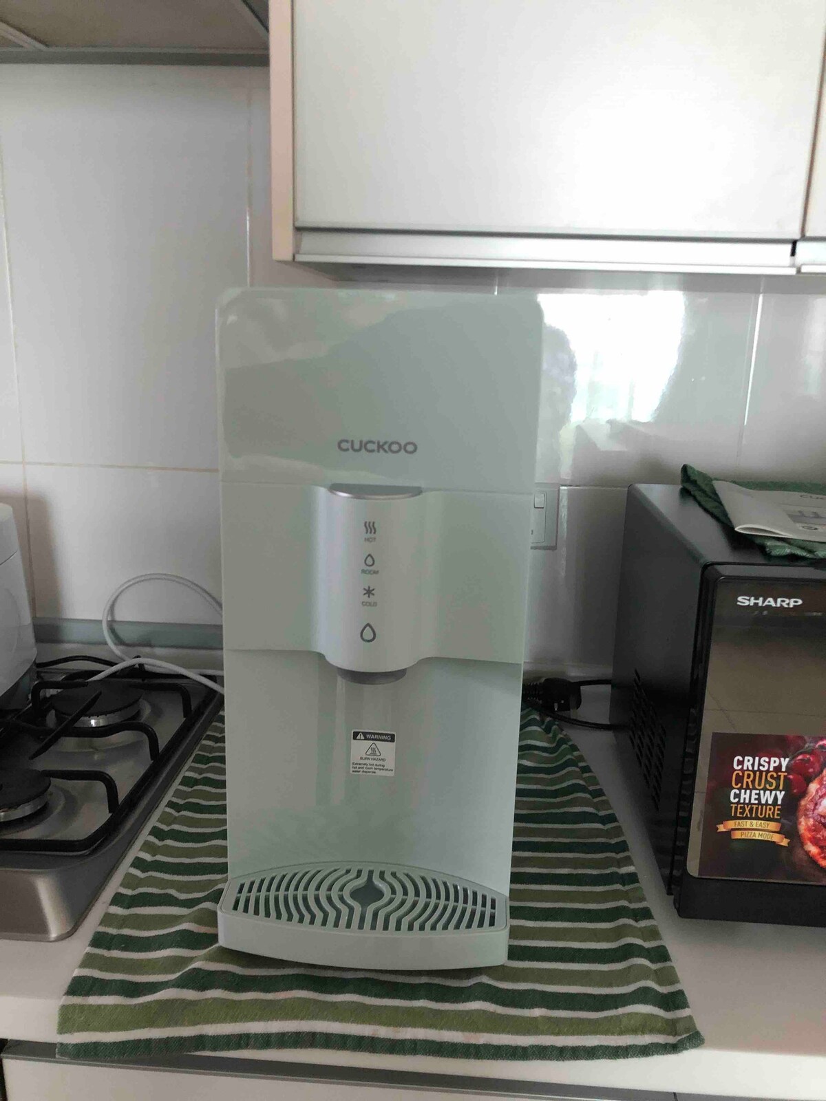 A modern water dispenser is positioned on a countertop, showcasing a sleek design in light green. It features user-friendly control buttons on the front. A patterned green and white dish towel is placed beneath it, while a stainless steel gas stove is visible in the background.