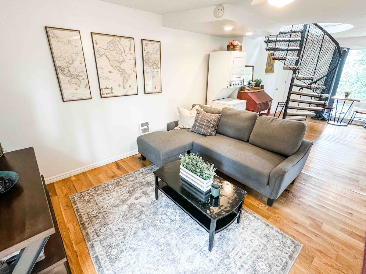 The living area features a gray sectional sofa positioned beside a black coffee table adorned with a plant and decorative items. Three vintage map prints hang on the wall, and warm hardwood flooring adds a cozy touch. A staircase leads to the upper level.
