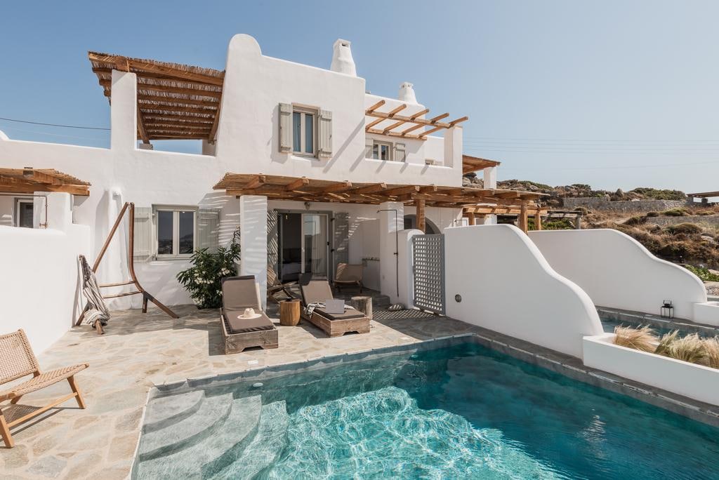 The exterior of the villa is shown, featuring a private pool with clear blue water and a stone deck. Sun loungers are positioned nearby under a wooden pergola. Whitewashed walls and large windows create a bright and airy atmosphere.