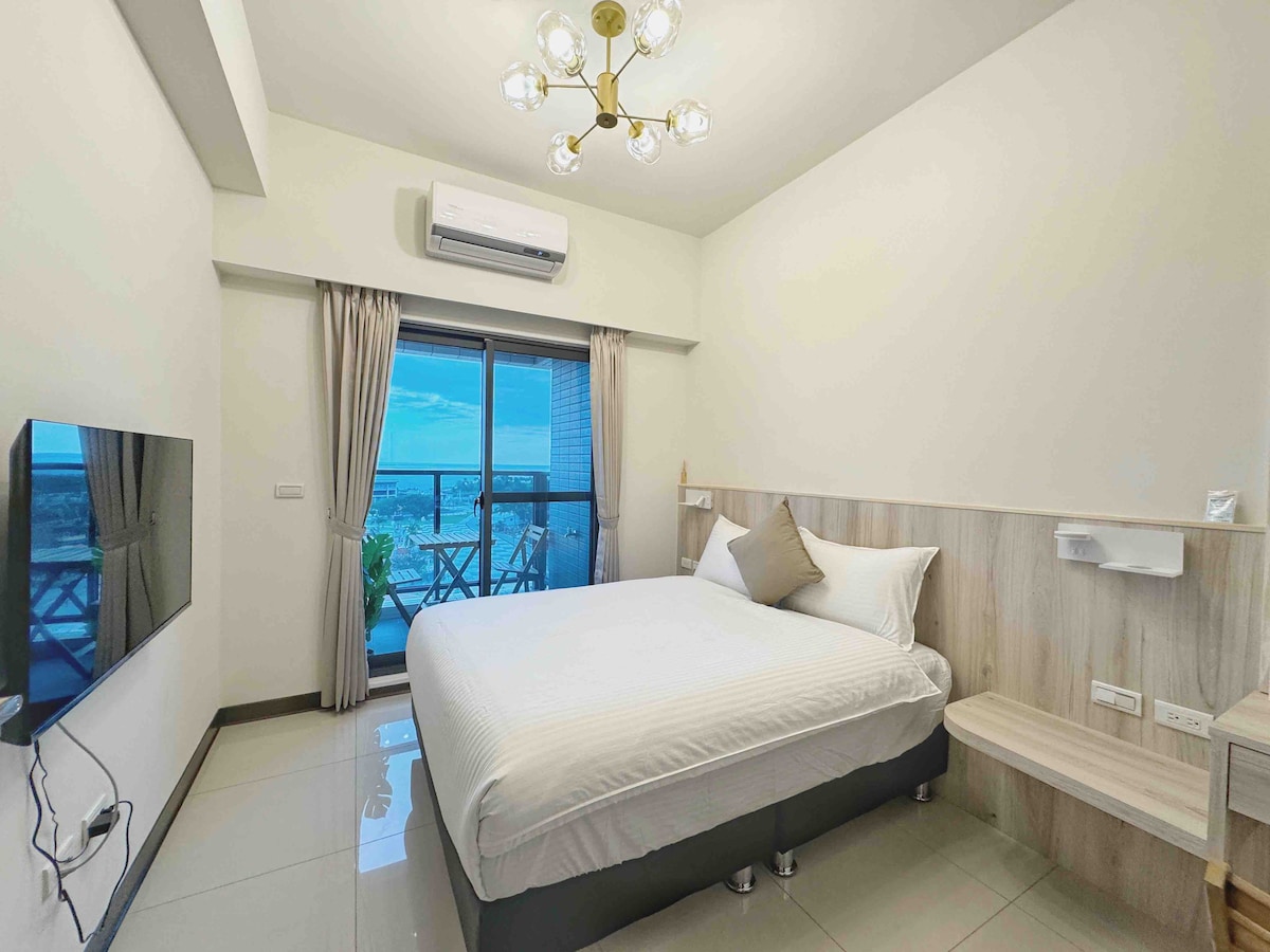 A bright bedroom features a spacious bed, light-coloured walls, and a large window that invites natural light. A flat-screen TV is mounted on one side, and a small shelf is attached nearby. A balcony door opens to views beyond, enhancing the room's airy feel.