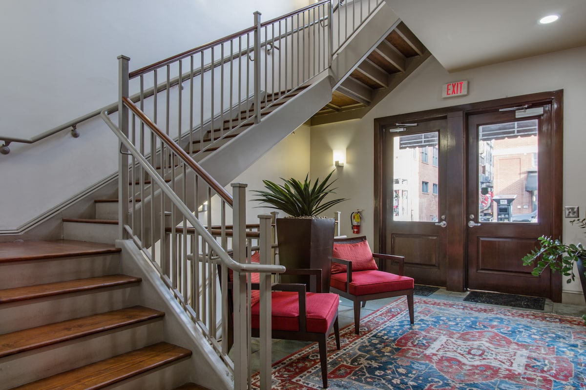 A welcoming entryway is showcased, featuring a staircase with metal railings and wooden steps. Two red chairs are positioned on a patterned area rug. A potted plant adds a touch of greenery to the space, which is complemented by wooden double doors leading to the outside.