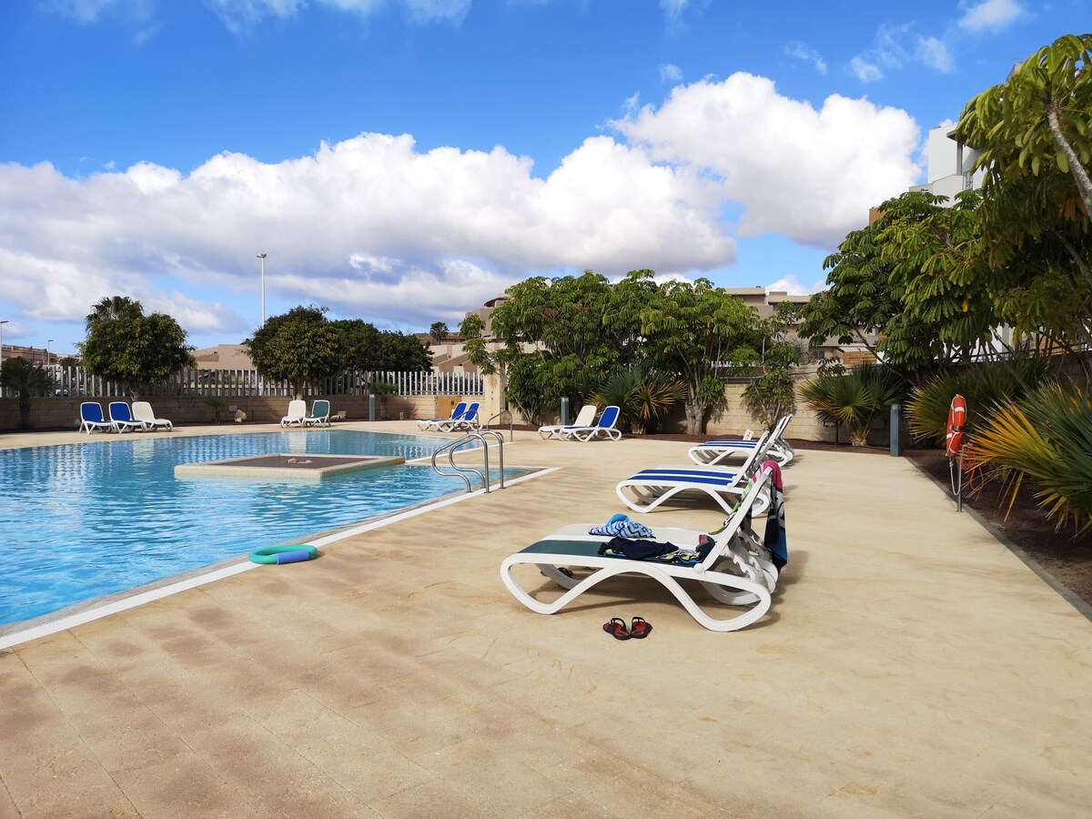 A sparkling pool is surrounded by a sunlit patio, with lounge chairs positioned for relaxation. Lush greenery adds a touch of nature to the scene. A colorful pool float is visible in the water, alongside a diving board. The sky is bright with few clouds.