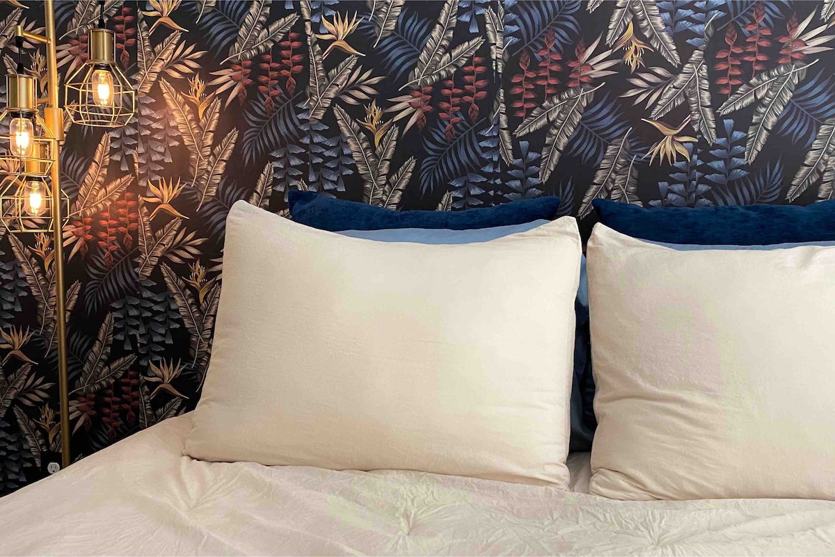 A well-appointed bedroom features a spacious bed with two large, soft pillows placed against a decorative wallpaper showcasing vibrant foliage patterns. Warm pendant lights hang beside the bed, adding a cozy touch to the inviting space.