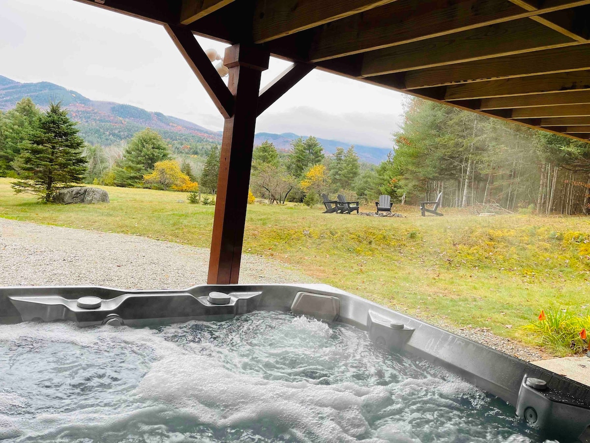 A private hot tub is situated beneath a wooden overhang, with bubbling water visible. In the background, rolling hills and trees create a natural setting, while a firepit area with arranged seating is discernible further afield.