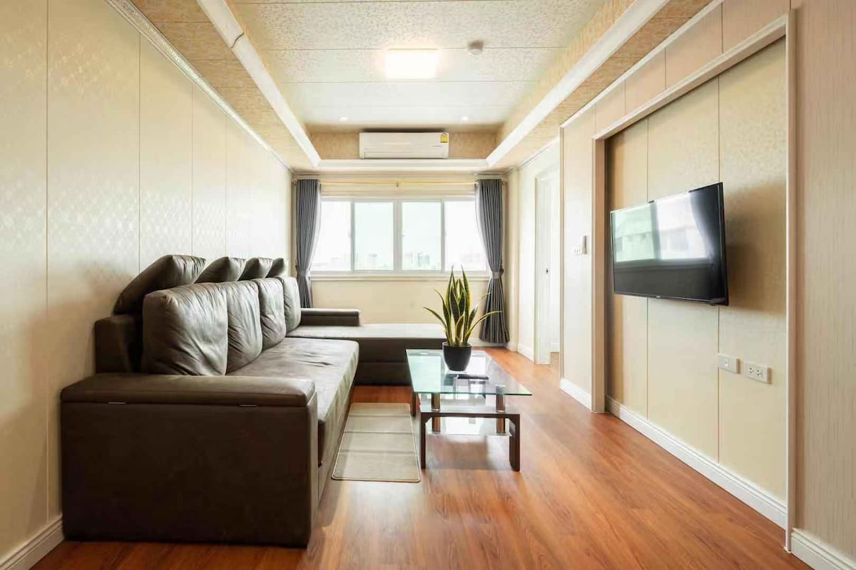 A modern living area features a large dark sectional sofa and a sleek glass coffee table. A flat-screen TV is mounted on the wall opposite a window allowing natural light to fill the room. Light-colored walls and wooden flooring enhance the spacious feel.