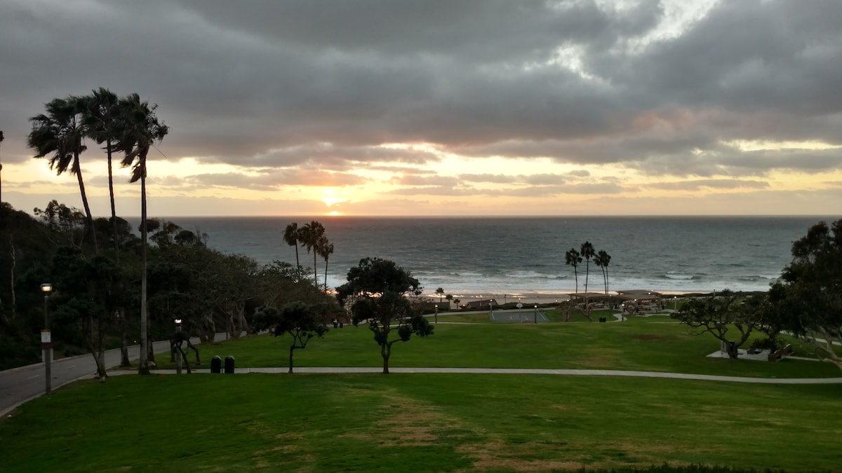 An expansive ocean view is displayed under a cloudy sky at sunset. The calm waves are visible alongside sandy shores, framed by palm trees and lush green park areas. A pathway along the beach runs parallel to the shoreline, enhancing the serene atmosphere.