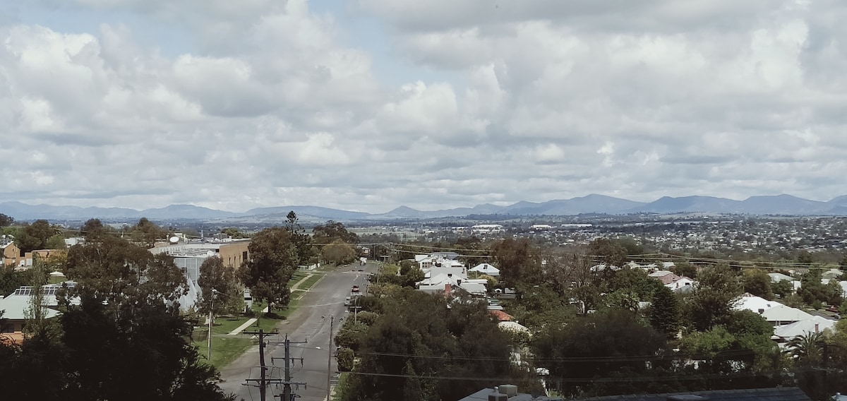 A panoramic view showcases distant mountains under a partly cloudy sky, with a peaceful neighborhood below. Trees line the streets, and residential homes are visible, emphasizing the serene and spacious setting of the area.