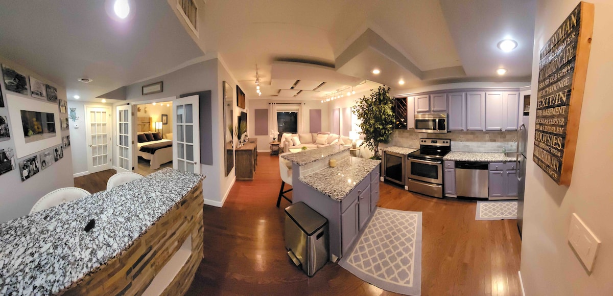 An open kitchen space features modern appliances, ample countertop space, and a breakfast bar with seating. Light-colored cabinets complement the granite surfaces. Adjacent areas showcase contrasting textures and artworks, while soft lighting enhances the welcoming atmosphere throughout the room.