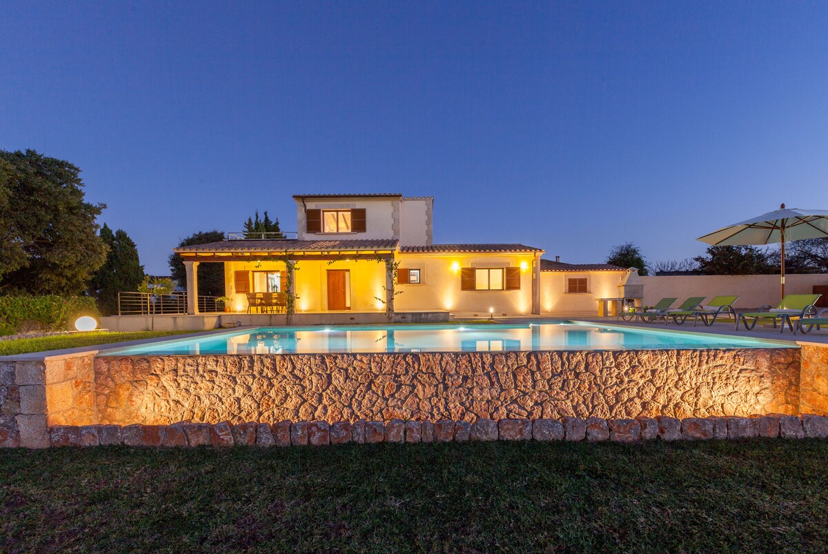 The villa is viewed from the pool area at twilight, showcasing its spacious two-story structure with warm lighting. An infinity pool reflects the surrounding gardens, while lounge chairs are neatly arranged nearby. The peaceful countryside setting enhances the tranquil ambiance.