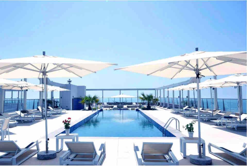 A rooftop pool area is shown under a clear blue sky, featuring a long swimming pool surrounded by lounge chairs with large umbrellas. Potted plants add greenery, while distant views of the sea can be seen, creating a serene environment.