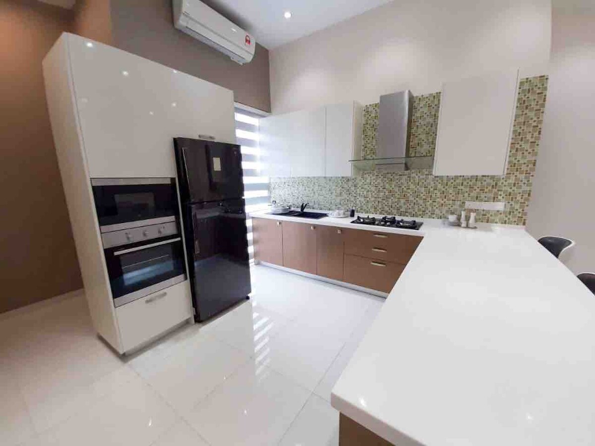 A modern kitchen features sleek cabinetry and a stylish backsplash with green tiles. Stainless steel appliances, including an oven and refrigerator, are present. A spacious countertop offers ample prep space, and natural light illuminates the area through window coverings.