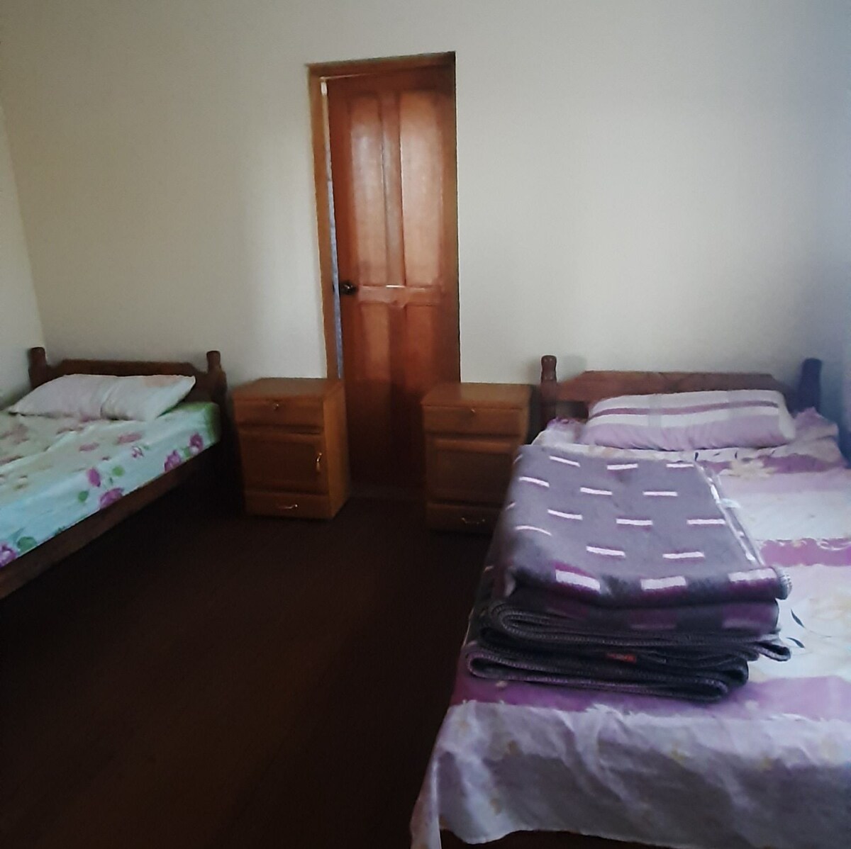 The room contains two beds with floral-patterned linens, positioned parallel to each other. A stack of blankets is neatly arranged on one bed. Two wooden nightstands sit beside each bed, while a wooden door leads to another area.