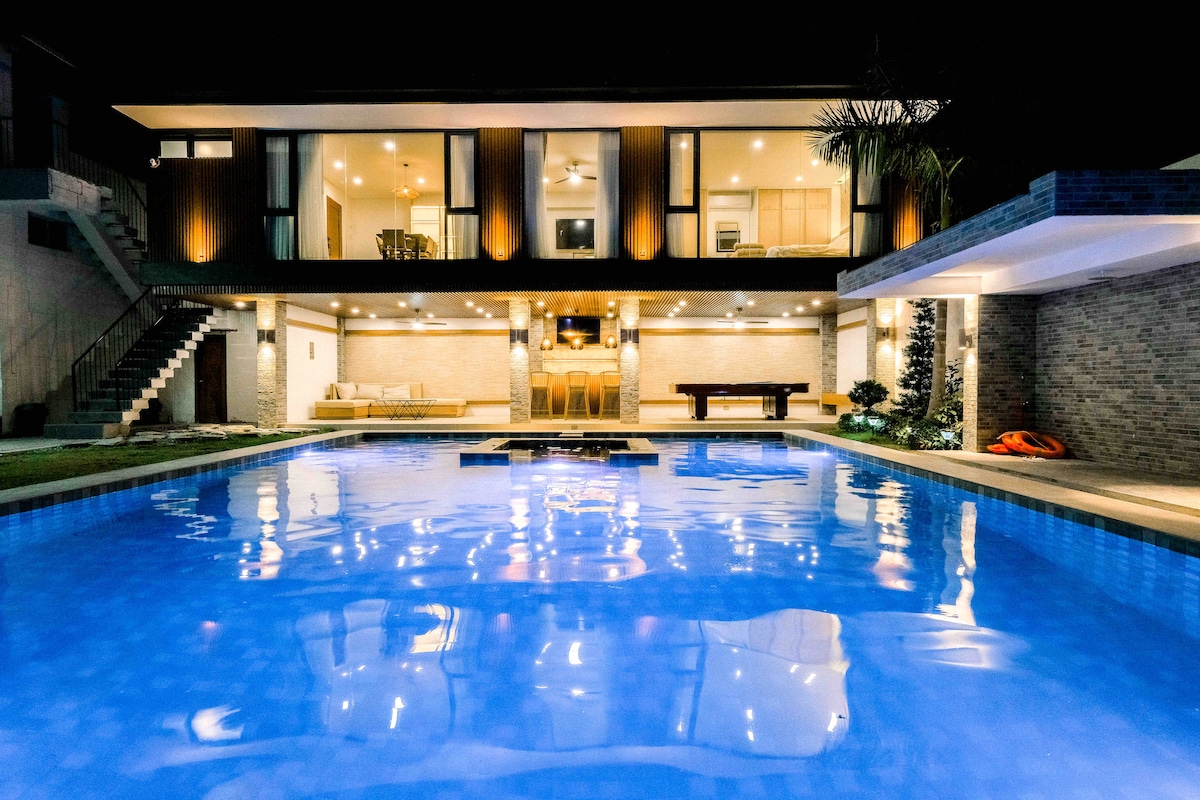 A large, illuminated swimming pool reflects the light from the surrounding outdoor area at night. The two-story building offers expansive glass doors leading to a spacious patio with seating and a billiard table. Lush greenery and comfortable lounge chairs are visible nearby.