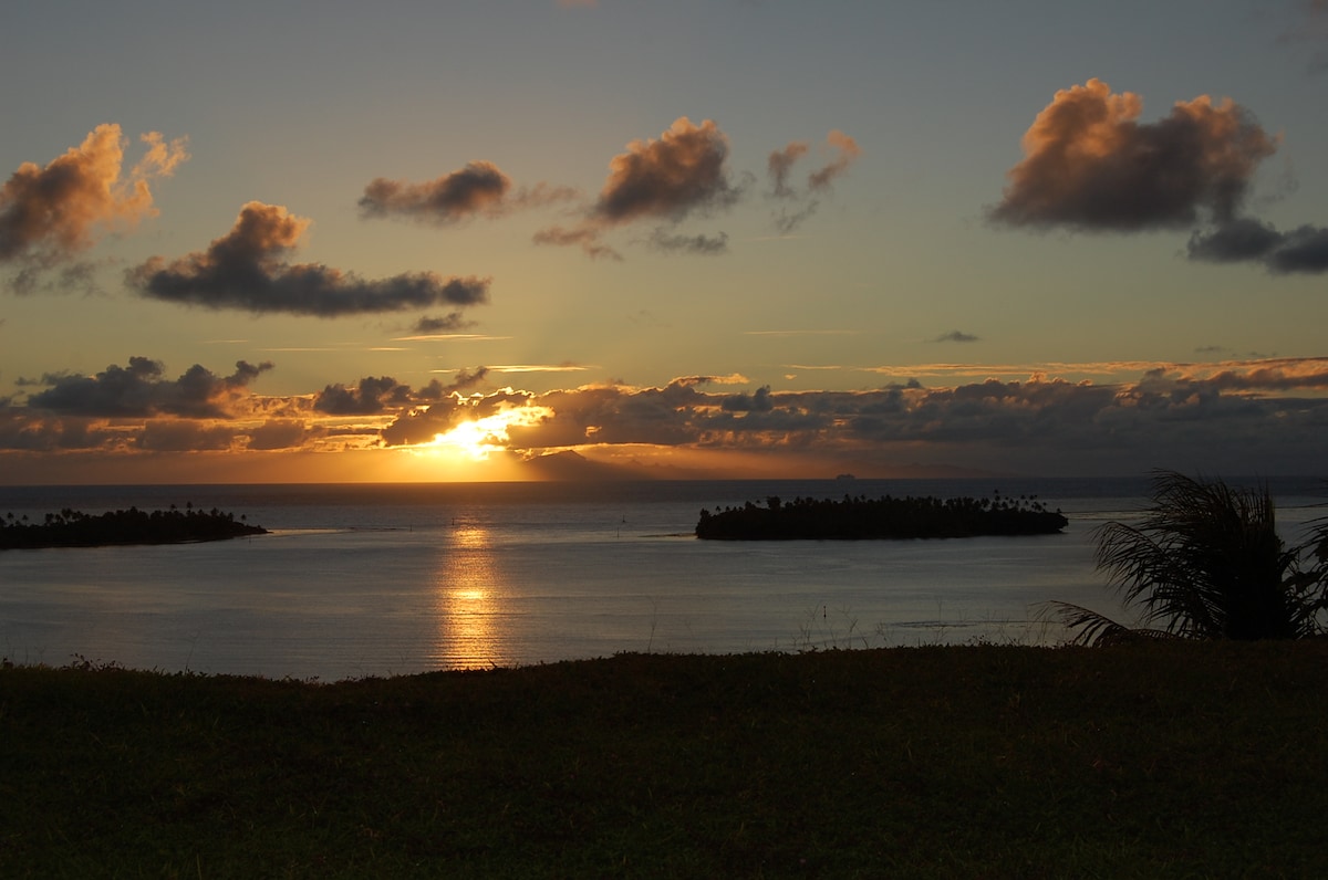 A serene sunset illuminates the sky with soft hues, casting a warm glow over the calm water. Lush land is framed in the foreground, while two small islands emerge from the tranquil lagoon, contributing to the natural beauty of the scene.