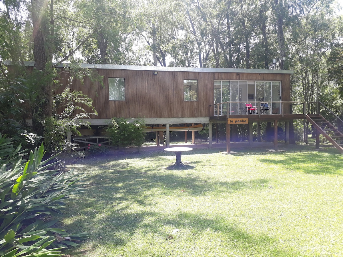 A modern wooden house is surrounded by greenery, elevated on stilts to create a unique design. Large windows allow natural light to fill the interior. A circular table sits on the well-maintained lawn in the foreground, providing a space for relaxation amidst the tranquil setting.