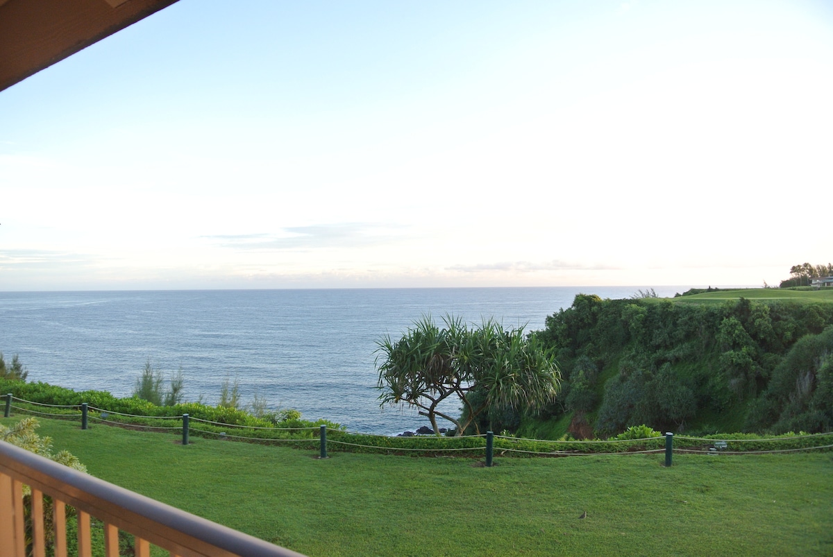 An expansive view of the ocean is captured from the balcony. Gentle greenery frames the scene, and a lone tree is positioned against the backdrop of the water. The sky is softly illuminated, transitioning into twilight.