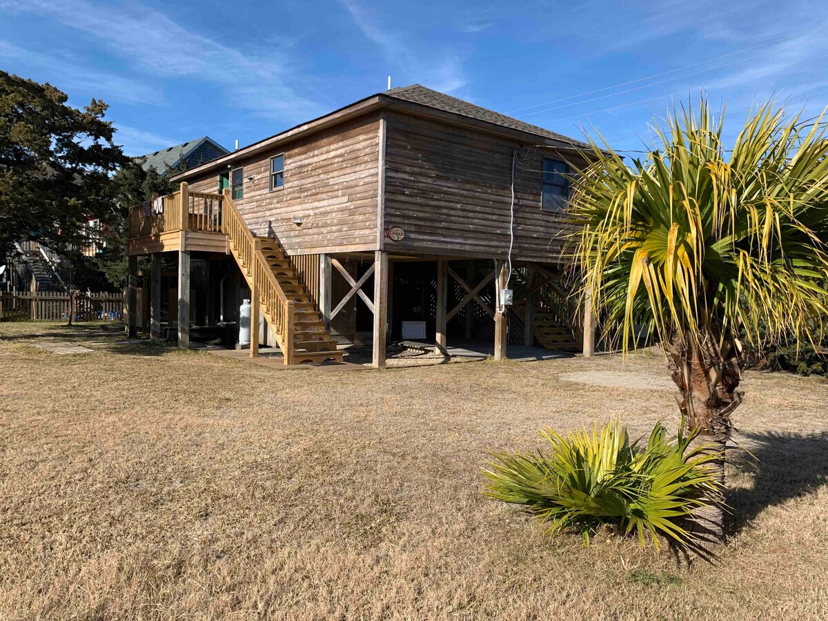 A wooden two-story home is showcased against a clear blue sky. Stairs lead up to the entrance, and a grassy area is visible in the foreground, accompanied by a palm tree. The house features a rustic appearance with horizontal wooden siding.