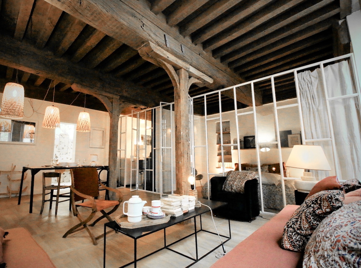An open living space features exposed wooden beams and a warm wooden floor. A cozy seating area is arranged with a sofa and armchairs, accompanied by a low coffee table stacked with books. The dining area is seen in the background, separated by glass partitions leading to a sleeping area.