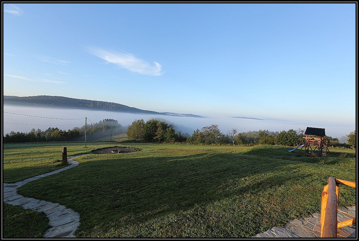 A serene landscape features rolling hills under a clear blue sky. A grassy area extends toward the horizon, where fog gently blankets the valley. A fire pit is visible in the foreground, along with a wooden play structure, enhancing the area’s natural beauty.