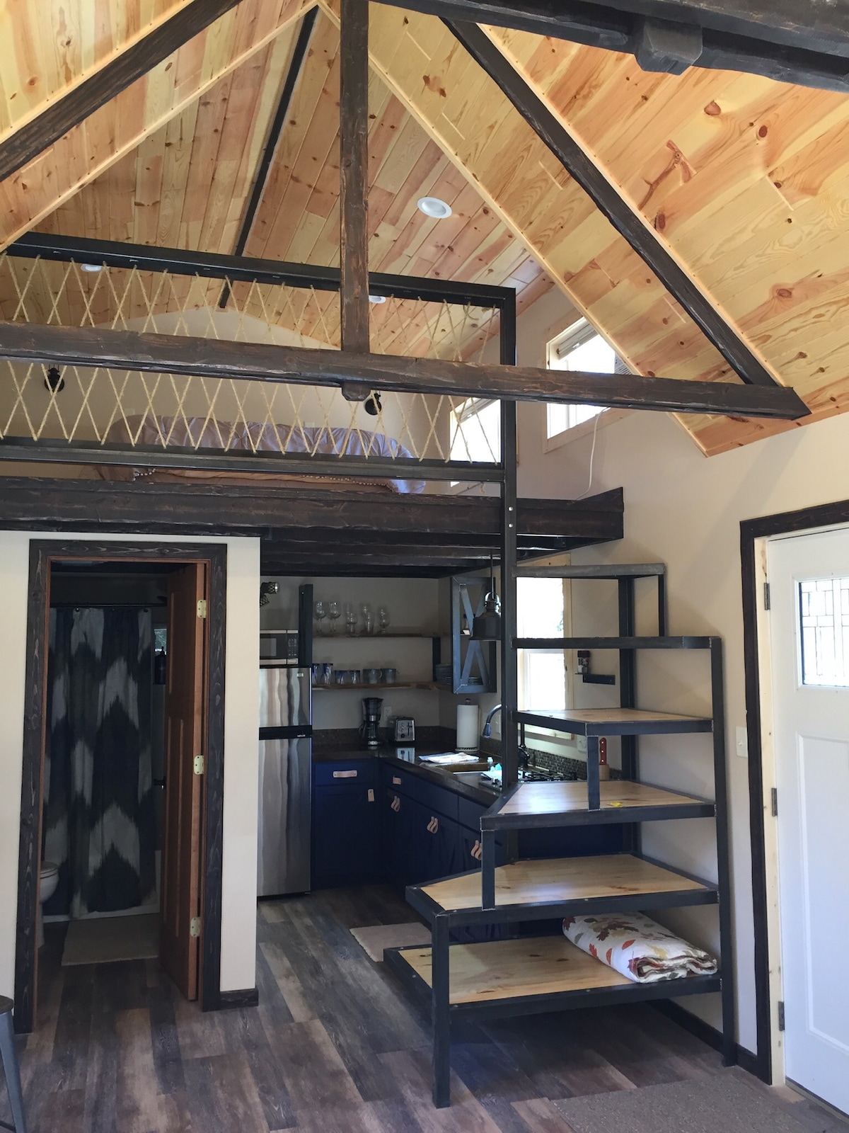 An open layout reveals a cozy interior with a loft area and a steep spiral staircase. The kitchen features dark cabinetry and modern appliances. The wooden ceiling adds warmth, and natural light filters through strategically placed windows.