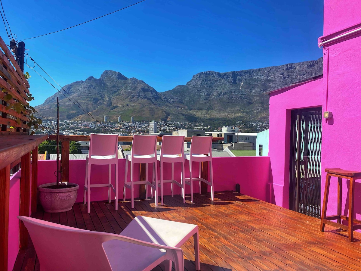 A private wooden deck is displayed, featuring high-backed bar stools overlooking a panoramic view of Table Mountain and the city below. The vibrant pink exterior of the property adds a pop of color against the clear blue sky, with sunlight illuminating the expansive outdoor space.