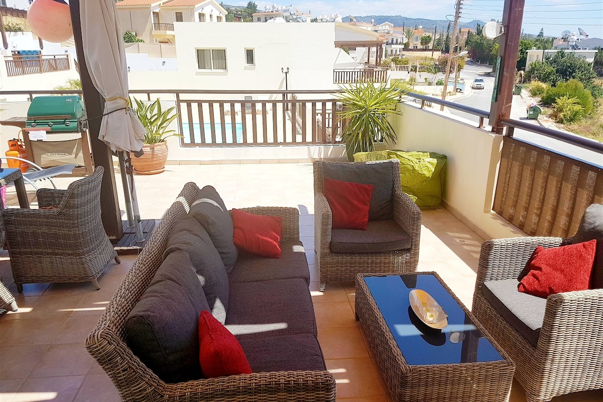 A spacious outdoor living area features comfortable rattan seating with vibrant cushions. A glass-top coffee table sits at the center, and the space is surrounded by views of the pool and distant hills. Sunlight fills the terrace, creating an inviting atmosphere.