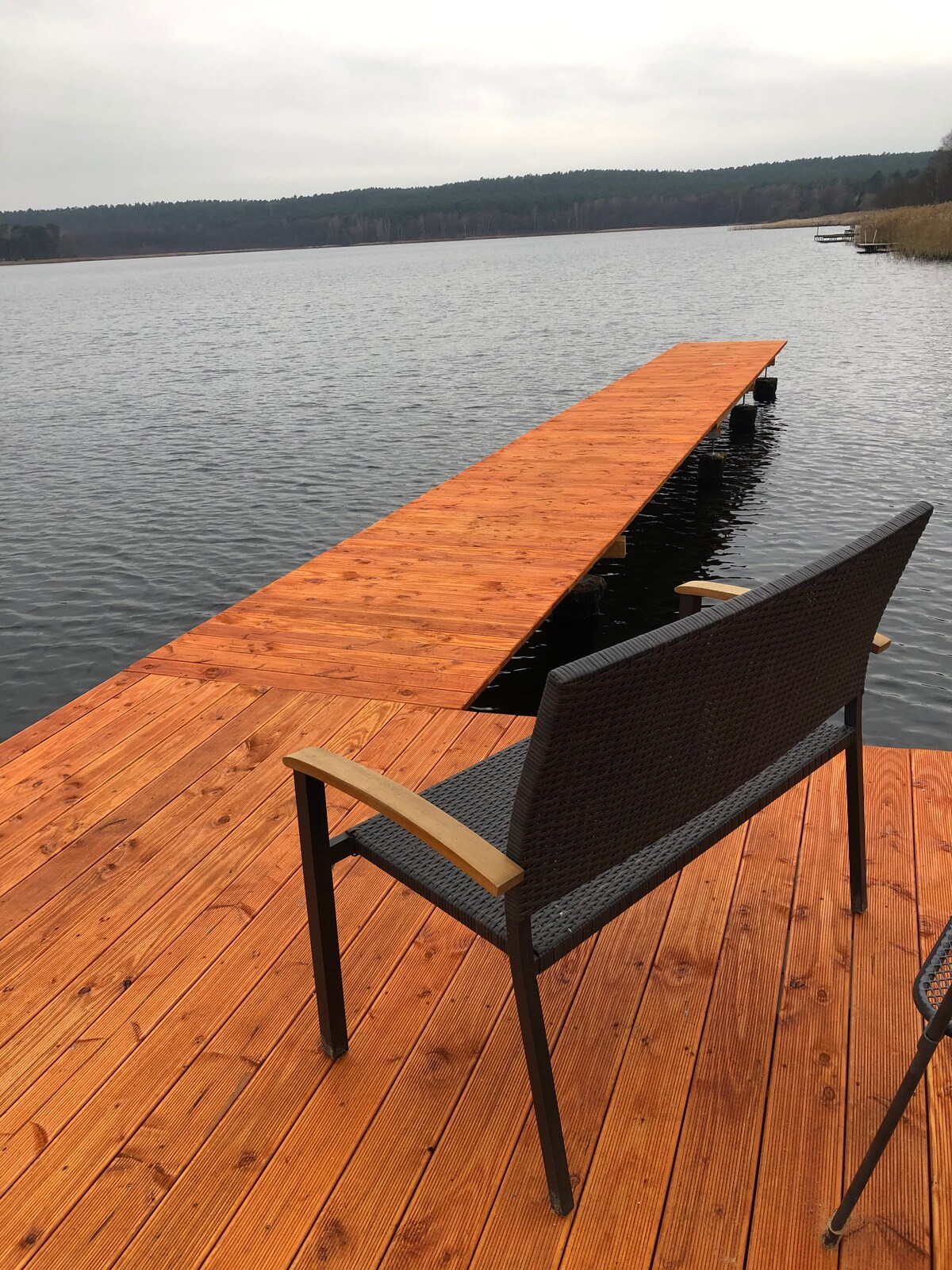 A wooden dock extends into the calm waters of the lake, with a single chair positioned on the edge. The wooden surface is polished, and the surrounding landscape features dense forest and gentle hills in the distance, providing a serene natural setting.