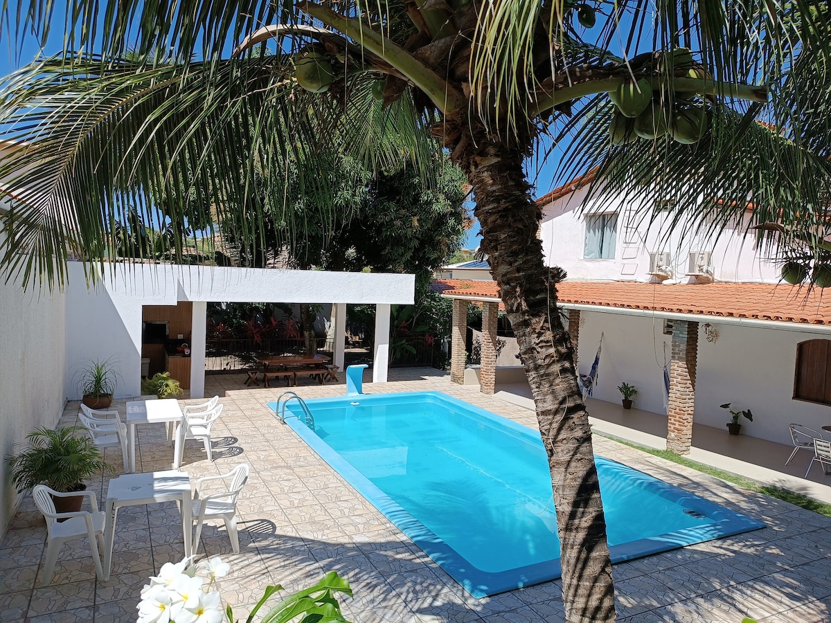 The image depicts a bright outdoor area featuring a large swimming pool surrounded by tiled flooring. Palm trees and other greenery add to the atmosphere. There are several white chairs and tables arranged nearby, with a shaded seating area in the background.