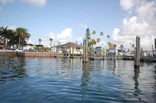The image captures a tranquil water view of the cottage from the bay, with calm waters reflecting the blue sky and fluffy clouds. Palm trees line the shore, and boats are anchored along the docks, providing a glimpse of the coastal lifestyle.