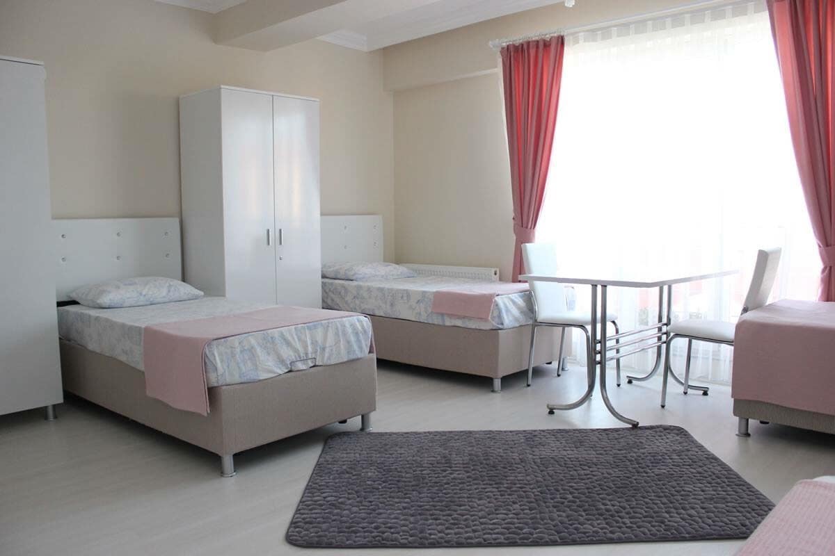 A spacious room is furnished with two beds, each dressed in light bedding and positioned next to a large window with sheer curtains. A round dining table is set with two chairs, and a light-colored rug lies on the floor.