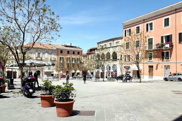 A lively city square features light-colored buildings with balconies adorned with plants. Elegant arches are visible in the facade, while well-maintained trees provide some shade. The pavement is paved with stones, and a few visitors can be seen walking through the area, enjoying the pleasant atmosphere.