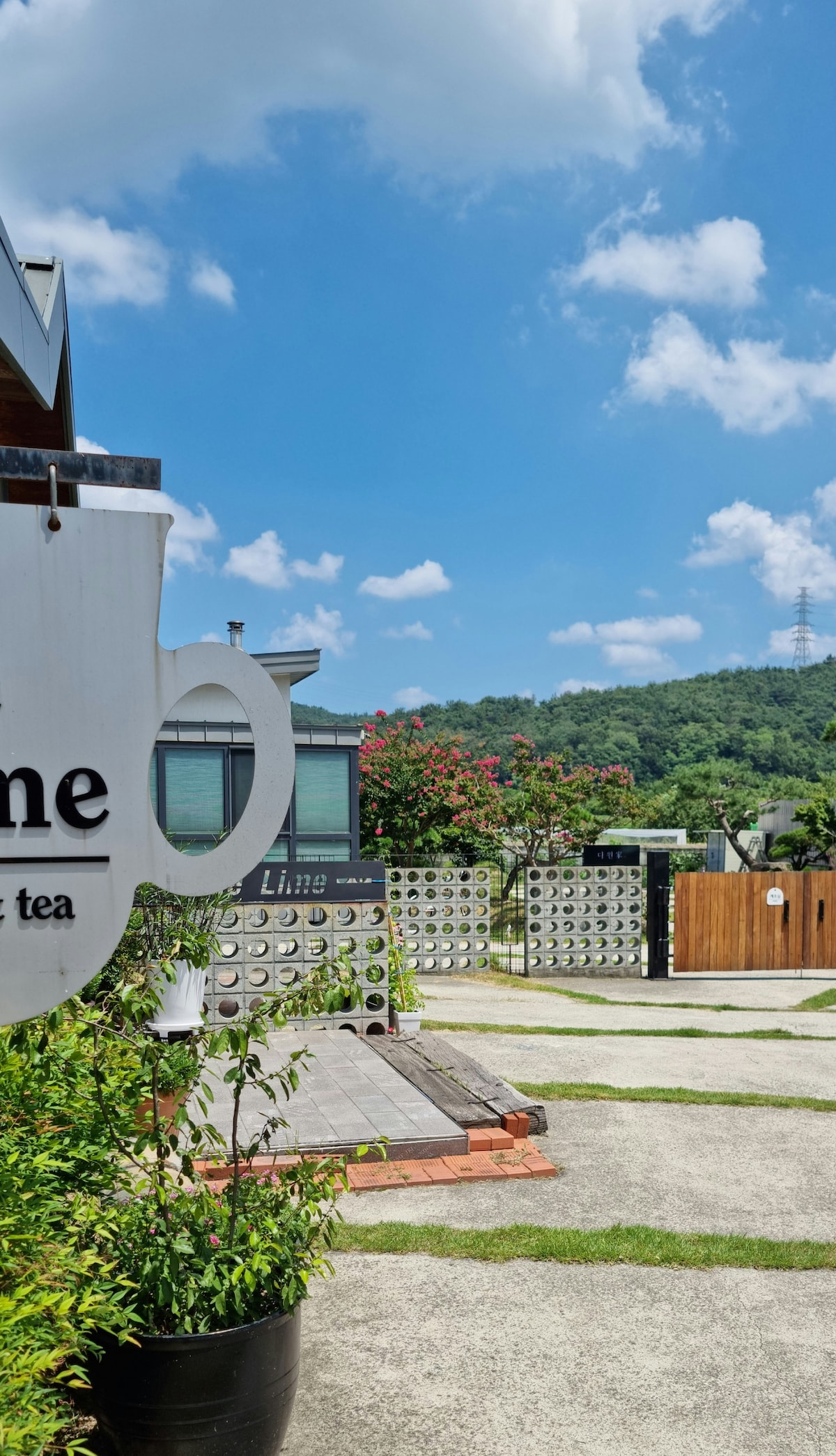 A serene outdoor area is shown, featuring a welcoming sign shaped like a coffee cup. Lush greenery and flowering trees are visible, enhancing the natural surroundings. A clear blue sky is present, with a distant hill and a power line adding context to the landscape.