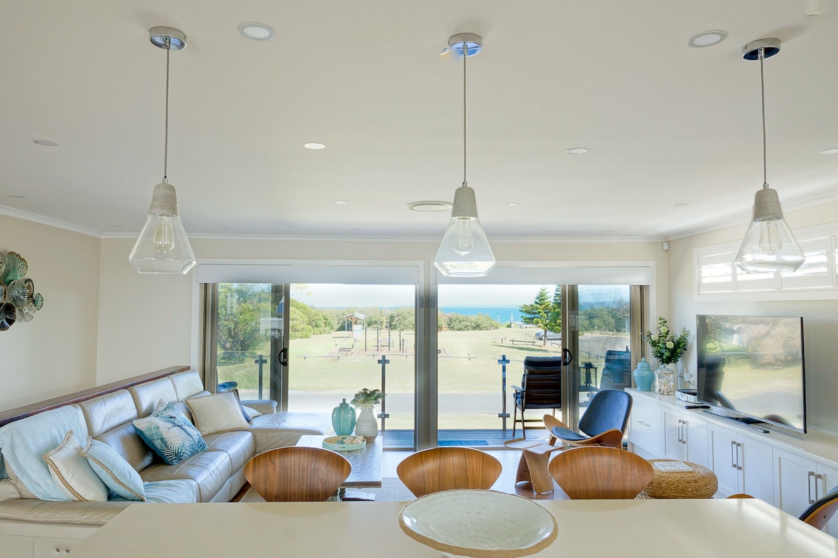 An open-plan living area showcases a comfortable seating arrangement with light-colored leather sofas and wooden chairs. Large sliding doors lead to a scenic view of the ocean, while pendant lights add subtle illumination. A flat-screen TV is positioned next to decorative elements, enhancing the space.