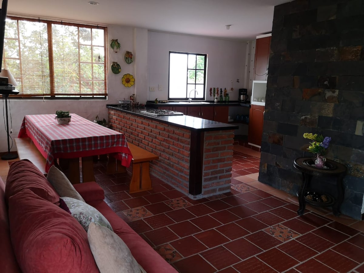 A bright kitchen and dining area features a large dining table with a red and white checkered tablecloth, complemented by wooden benches. The kitchen is equipped with a granite countertop and modern appliances. Natural light fills the space through large windows, highlighting the warm tones of the brick floor.
