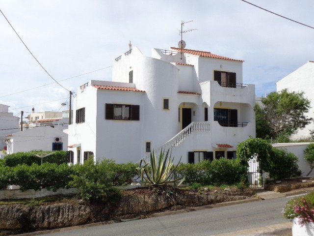 A white, multi-level building features traditional Mediterranean architecture with a sloped red tile roof. Windows with dark frames and a decorative balcony are visible, enhancing the structure's charm. Lush greenery lines the property, contrasting with the light exterior and creating a welcoming appearance.