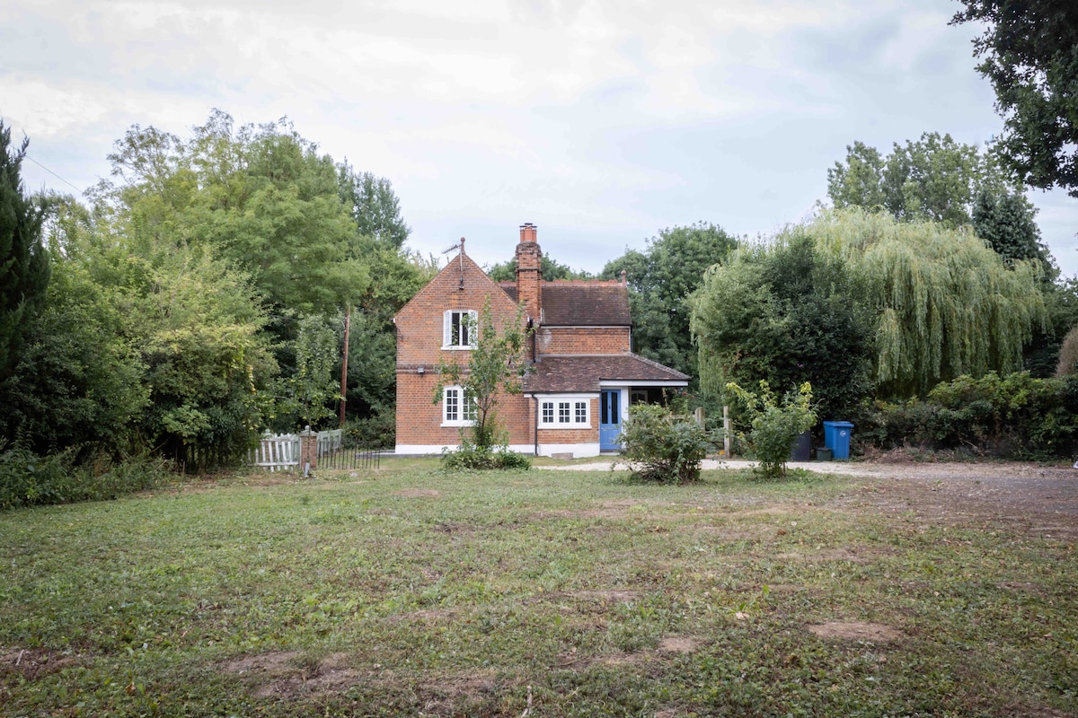 The cottage features a traditional brick exterior with a blue front door, surrounded by a natural landscape of trees and shrubs. A spacious area of lawn is visible in front, offering a sense of privacy and tranquility.