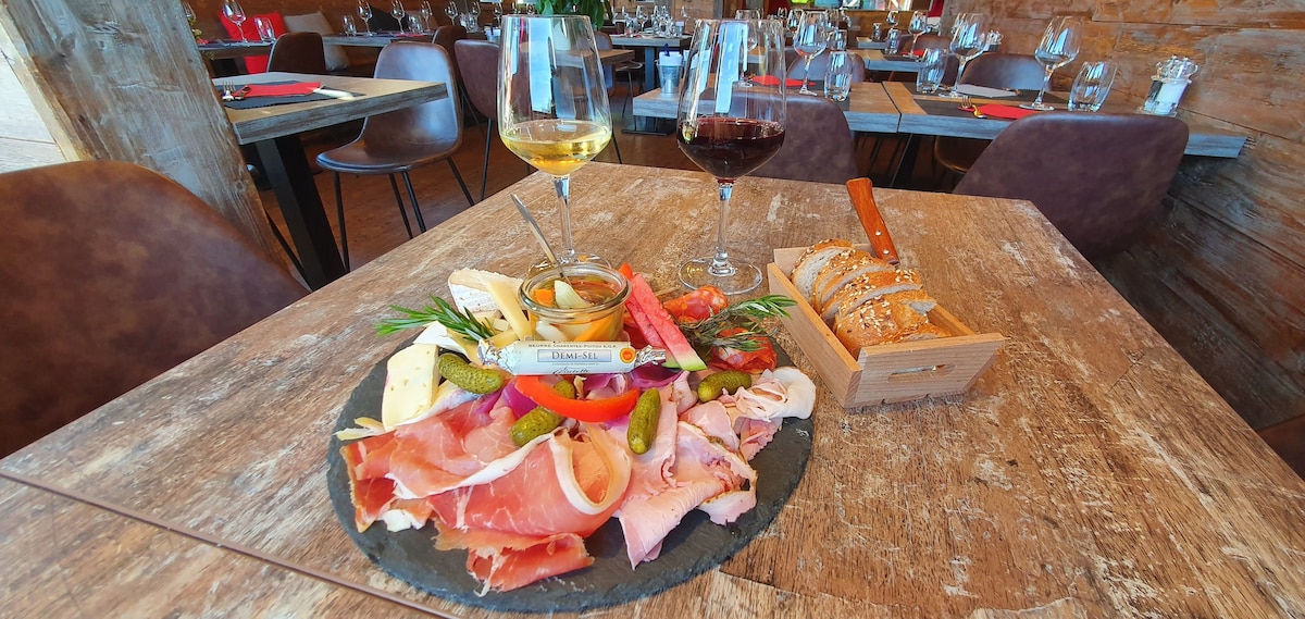 A wooden dining table is set with a variety of cured meats, cheeses, olives, and colorful vegetables arranged on a slate platter. Two glasses of wine, one white and one red, sit beside the platter. The background features a rustic dining area with multiple tables and chairs.