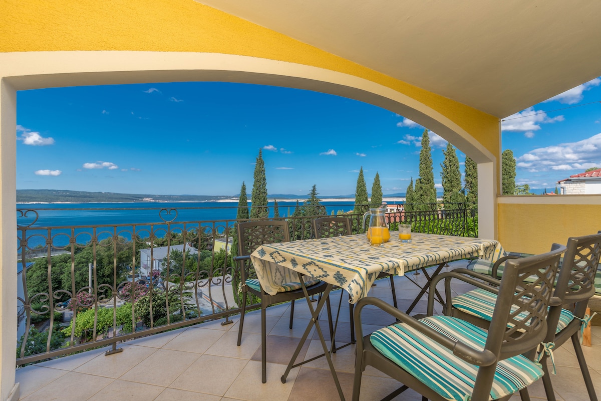 A spacious terrace is presented with a patterned tablecloth and several chairs, offering a view of the sea and surrounding landscape. Lush greenery is visible in the background, along with a clear blue sky.