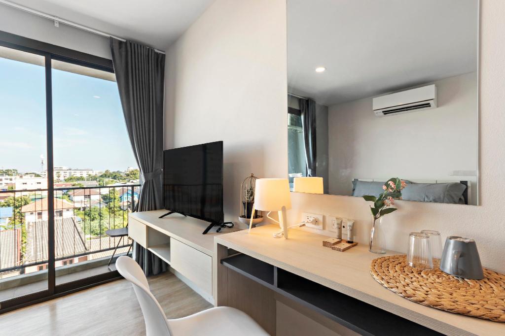 A comfortable workspace is created with a wooden desk and a white chair, positioned in front of a flat-screen TV. Natural light enters through sliding glass doors, leading to a balcony. A small decorative lamp and a minimalist arrangement of glasses are displayed on the desk.