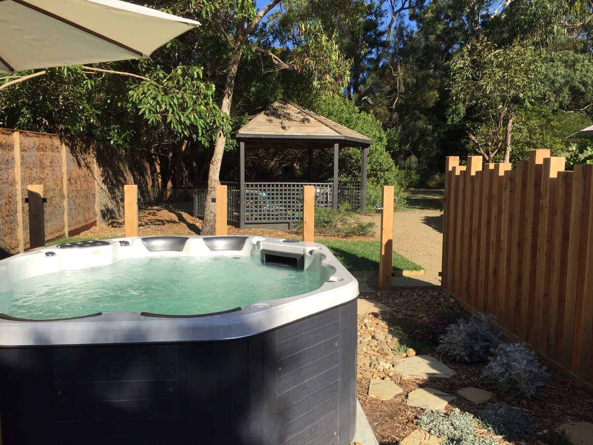 A safety-fenced spa is positioned within a rustic garden. Sunlight reflects off the water surface, while a gazebo is visible in the background. Surrounding greenery provides privacy and a serene atmosphere, with a wooden fence marking the property boundary.
