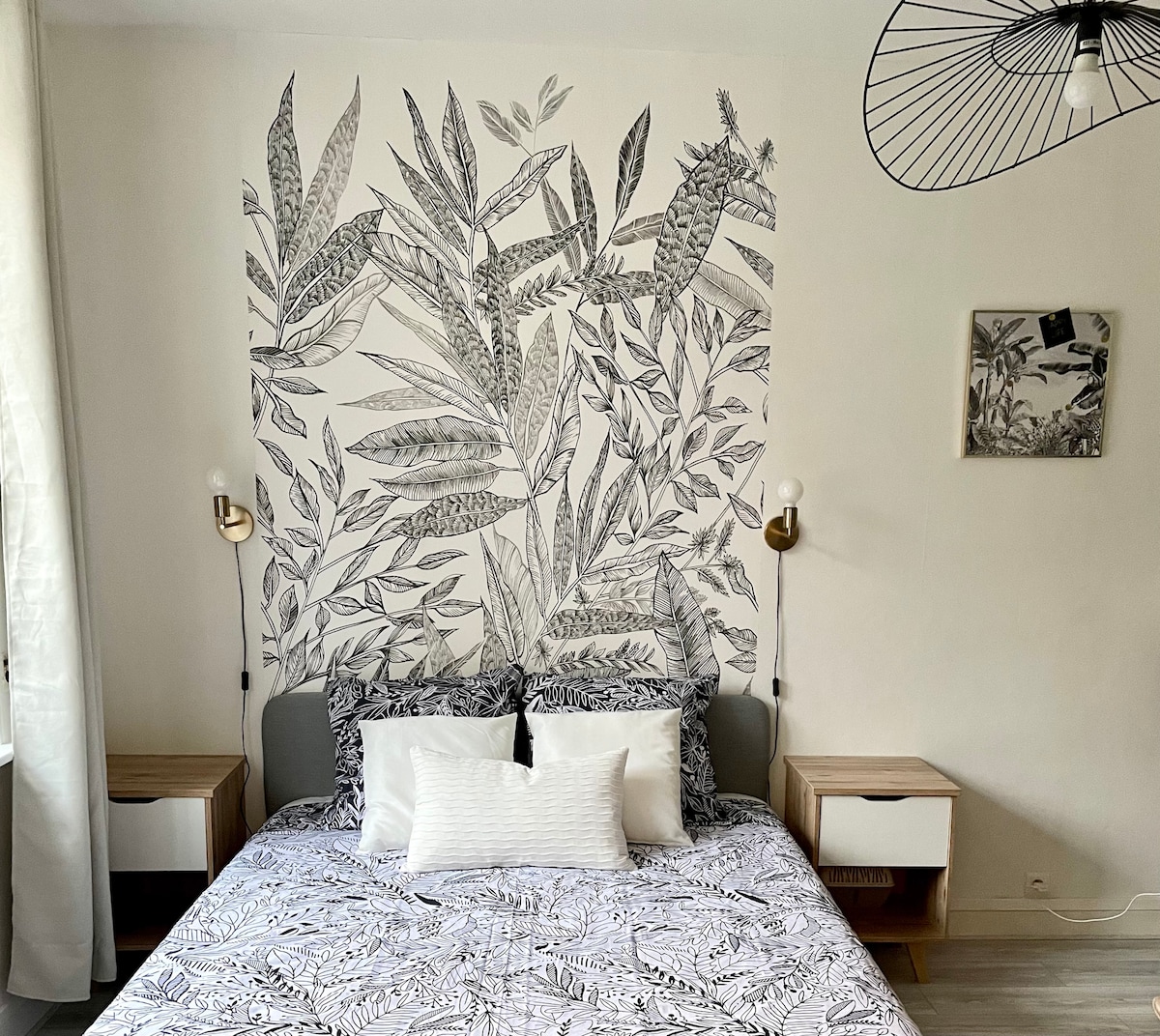 A comfortable sleeping area features a spacious bed with patterned bedding, flanked by two wooden nightstands. A nature-inspired wall mural adds a touch of elegance. Soft light is provided by modern wall sconces, with natural light filtering through sheer curtains.