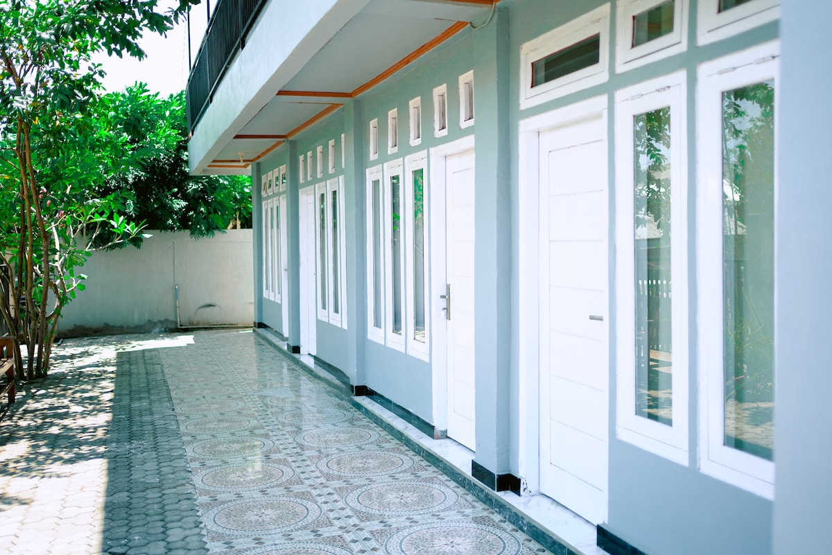 An exterior view of the guest house features a tiled pathway adorned with greenery. Several rooms are visible along the side, each with white doors and large windows allowing natural light. The simple design emphasizes a clean and functional aesthetic.