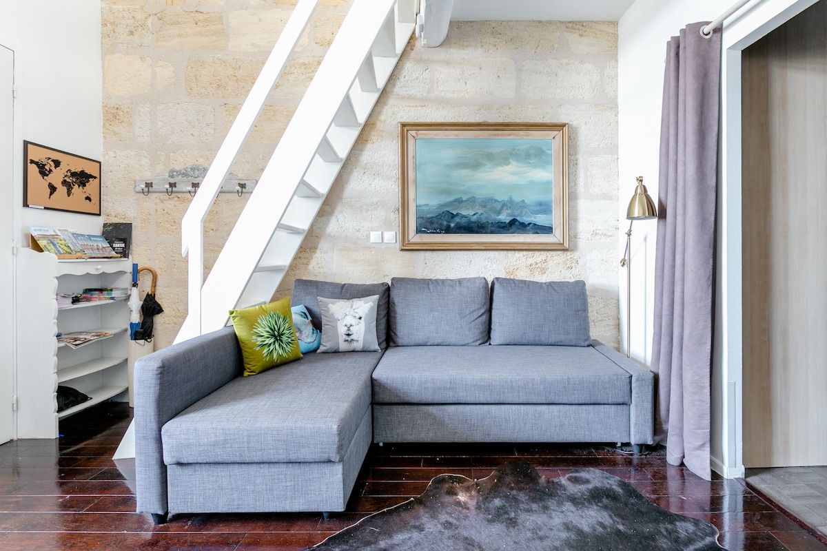 A modern gray sectional sofa occupies one corner of the living area, accompanied by a decorative pillow and a small leather rug beneath it. A framed landscape painting adorns the wall above the sofa. A white staircase leads to the upper level, complementing the exposed stone walls.