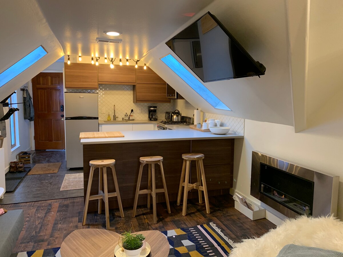The modern kitchen features sleek cabinets and an island with three wooden stools. Under-cabinet lighting illuminates the workspace. A seating area is visible, complemented by a cozy fireplace and a rug with geometric patterns. Skylights provide natural light throughout the space.