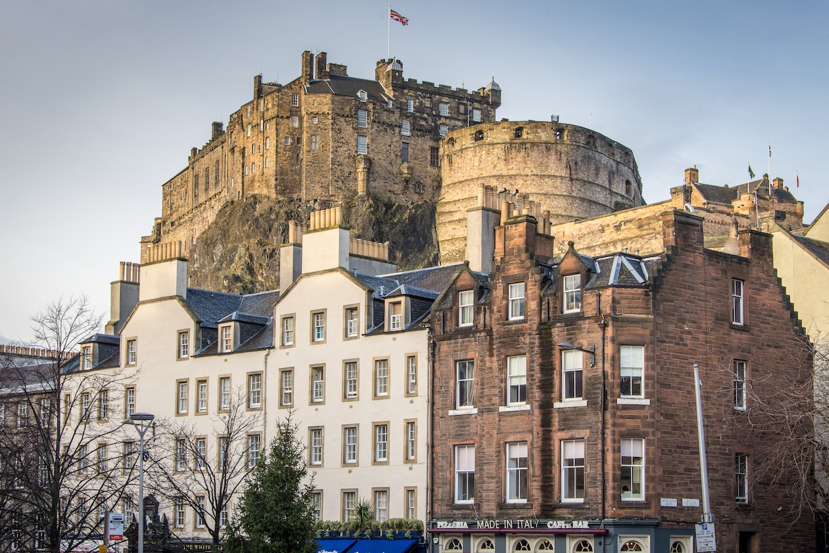 The image showcases Edinburgh Castle perched atop Castle Rock, displaying its historic stone structure and iconic towers. Below, a row of classic buildings features a mix of architectural styles, with a clear view of well-preserved facades and windows, set against a bright sky.