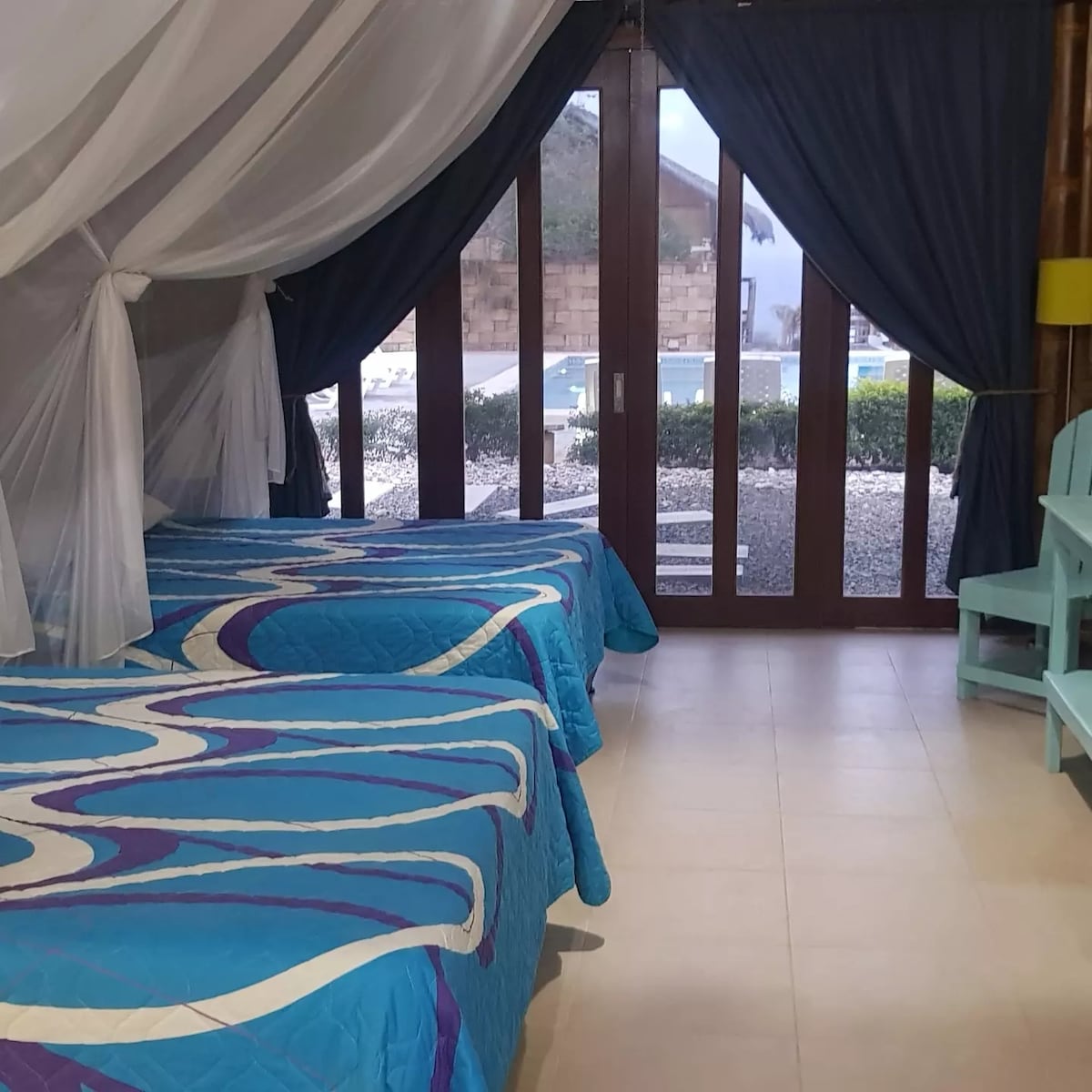 The room features two spacious beds draped with light, airy canopies. Large glass doors provide a view of the outdoor area, which includes a pool. Soft blue bed linens with wavy patterns complement the serene atmosphere, while dark curtains add depth to the room.