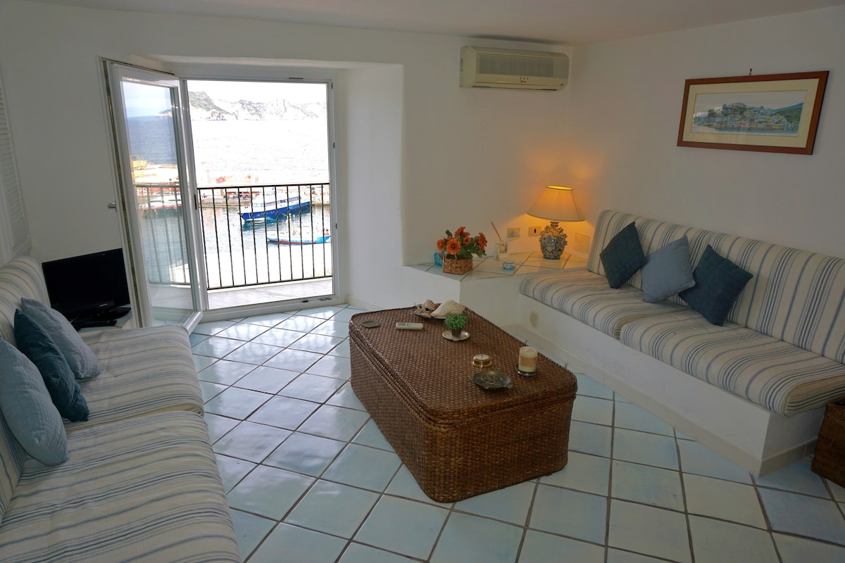 A bright and airy living area features two striped sofas and a wicker coffee table, emphasizing a relaxed atmosphere. Natural light floods the space through an open glass door that leads to a balcony overlooking the harbor, with boats visible in the background.
