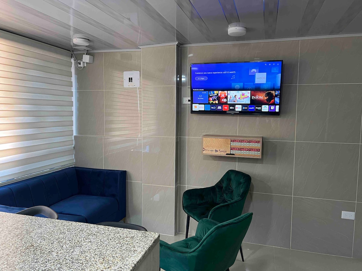A modern living area features a wall-mounted smart TV above a stylish media console. Two armchairs, one in green velvet and another in warm tones, complement a cozy corner sofa. Soft natural light filters through layered window shades, enhancing the sleek design of the space.