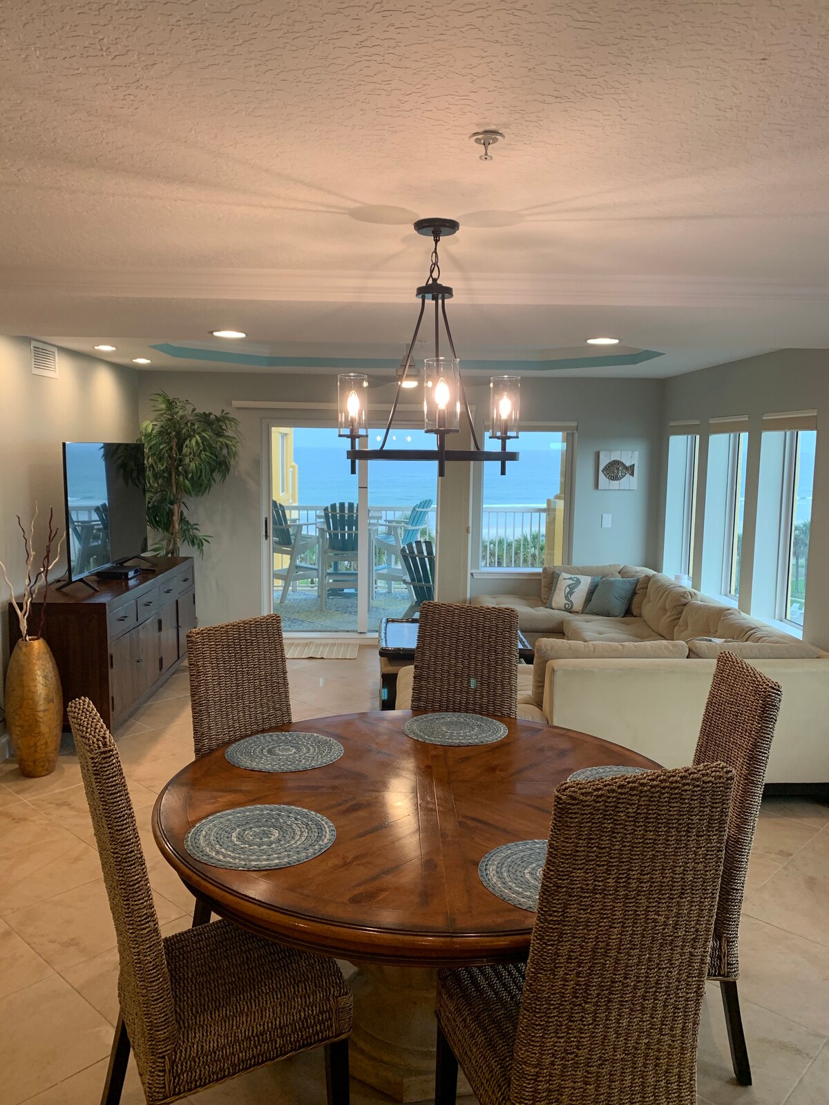 A dining table with four woven chairs is positioned in the foreground, showcasing a natural wood finish. In the background, a spacious living area is visible, featuring large windows that offer views of the ocean. A light fixture hangs from the ceiling, illuminating the area.